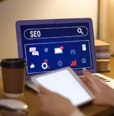 A close-up of a person holding a tablet while a laptop in the background displays a search bar with the text "SEO" above a panel of digital marketing icons, with a coffee cup nearby